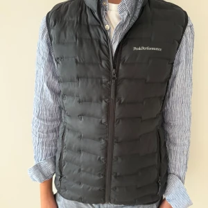 Svart dunväst från Peak Performance - Snygg svart dunväst från Peak Performance med quiltad design och hög krage. Västen har dragkedja framtill och två praktiska fickor med dragkedja. Perfekt att slänga över en hoodie eller skjorta för en chill look. Nypris ca 1000kr, hör av er vid frågor! Passformen är lite liten.