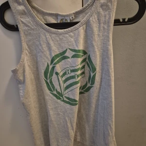 Grå Hammarby IF linnetopp - Snygg grå ärmlös linnetopp från Hammarby IF med klubbens klassiska gröna logga och flagga framtill. Perfekt för dig som vill visa ditt stöd på matchen eller bara gillar sportig stil. Mjuk bomullskänsla och avslappnad passform.