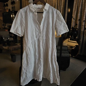 Vit kort klänning i broderad bomull - Superfin vit kortklänning från Cubus i broderad bomull med hålmönster. Klänningen har korta ärmar, knappar framtill och klassisk krage. Perfekt för varma dagar med sin luftiga känsla och snygga detaljer.