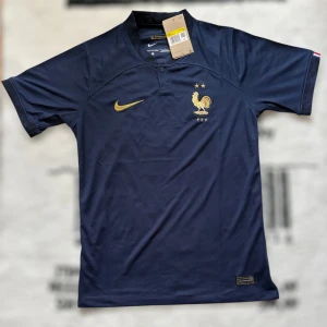 Frankrike fotbollströja marinblå - Marinblå officiell Frankrike fotbollströja från Nike med guldiga detaljer, broderad tupp och två stjärnor på bröstet. Tröjan har korta ärmar, liten krage med knapp och är tillverkad i lätt, ventilerande funktionsmaterial. Perfekt för dig som älskar landslagsfotboll.