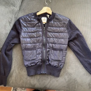 Mörkblå moncler liknande jacka från Gina tricot  - Snygg mörkblå jacka från Gina Tricot med quiltad framsida och två fickor. Ärmar och rygg är stickade för en soft känsla, medan framsidan är i glansigt material. Dragkedja framtill och ribbade muddar ger en clean look.