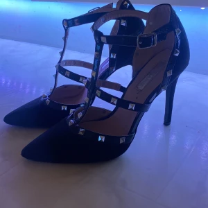 Svarta pumps med nitar och hög klack - Svarta pumps med spetsig tå och höga klackar. Skorna har flera remmar med silvriga nitar och spänne runt vristen. Snygg och edgy look som sticker ut. Perfekta för dig som gillar detaljer och vill ha en statement-sko.