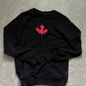 Svart Dsquared2 sweatshirt med lönnlöv - Svart sweatshirt från Dsquared2 med rött lönnlöv och broderad logga på bröstet. Tröjan har rund hals, ribbade muddar och är tillverkad i mjuk bomull. Perfekt för dig som gillar streetwear och vill sticka ut med en ikonisk detalj.