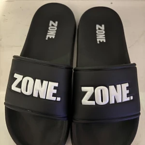 Svarta slides från ZONE strl 37/38 - Svarta slides från ZONE med vit logga på ovandelen. Skorna har en bred rem över foten och platt sula med mönstrad undersida för extra grepp. Tillverkade i syntetmaterial och perfekta för chill dagar eller efter träning. Köpta på klubbhuset. Aldrig använt pga fel storlek 