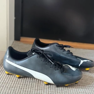 Svarta fotbollsskor från Puma - Svarta fotbollsskor från Puma med vit logga på sidan och framtill. Skorna har snörning och gula dobbar. Använda en gång. 