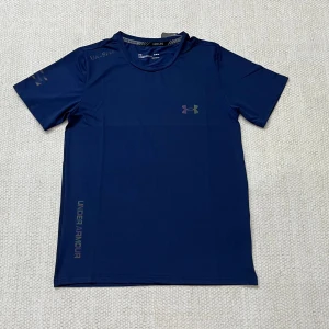 Marinblå träningst-shirt Under Armour - Sportig marinblå t-shirt från Under Armour med korta ärmar och diskret logga på bröstet. Tillverkad i lätt och snabbtorkande polyester, perfekt för träning. Snyggt tryck längs sidan och bekväm passform som funkar till alla aktiva dagar.