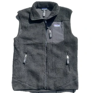 Svart fleeceväst från Patagonia - Mysig svart fleeceväst från Patagonia med hög krage och tre dragkedjeförsedda fickor. Västen har en klassisk, enkel design och är perfekt för lager-på-lager. Materialet är mjukt och fluffigt, och bröstfickan har en snygg Patagonia-logga.