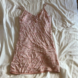 Rosa blommig kortklänning med knyt - Superfin rosa kortklänning med vita och gula blommor, smala axelband och söt knytning framtill. Klänningen har en lätt och flowy passform med dragkedja på sidan, perfekt för varma dagar. Materialet känns mjukt och luftigt, och mönstret ger en somrig vibe.