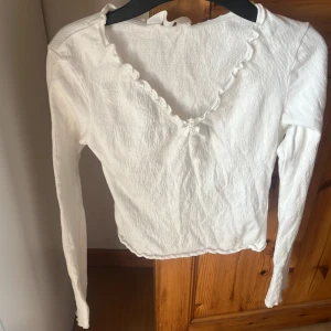 Vit långärmad blus med rosett H&M - Supersöt vit långärmad blus från H&M med v-ringning och liten rosett framtill. Blusen har vågig kant vid halsen och är gjord i ett lätt, strukturerat bomullstyg. Perfekt för dig som gillar en feminin och enkel stil.