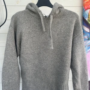 Grå stickad hoodie med huva - Mysig grå stickad hoodie med huva och snörning. Tröjan har långa ärmar och bred ribbad kant nedtill och vid ärmsluten. Perfekt för kyliga dagar när du vill vara både bekväm och snygg.