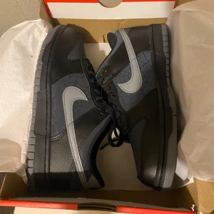 Nike Dunk Low svart/grå med spindelnätsmönster - Nike Dunk Low sneakers i svart och mörkgrått med coolt spindelnätsmönster på panelerna. Skorna har en ljusgrå swoosh, svarta skosnören och platt sula. Perfekta för dig som vill sticka ut med en unik detalj på dina kicks.