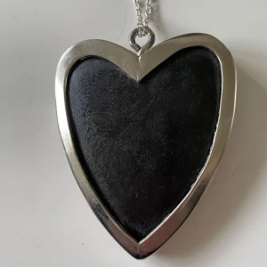 Halsband med stort  svart hjärthänge - Stilrent halsband med ett stort hjärtformat hänge i svart och silver, 6*8 cm. Hänget har en slät svart yta omgiven av en bred silverram och hänger i en tunn silverfärgad kedja. Perfekt för dig som gillar statement-smycken med enkel design.