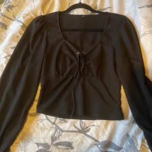 Svart croppad blus med snörning - Snygg svart croppad blus med långa puffärmar och knappar vid ärmsluten. Toppen har v-ringning med snörningsdetalj framtill och är gjord i ett lätt strukturerat tyg. Perfekt för dig som gillar trendiga och stilrena plagg.