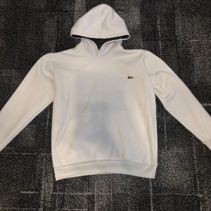 Vit hoodie från Lacoste med huva - Snygg vit hoodie från Lacoste med klassisk krokodil-logga på bröstet. Tröjan har huva, lång ärm och en stor magficka framtill. Perfekt för en chill och stilren look. Mjuk och skön känsla tack vare bomullsmaterialet.