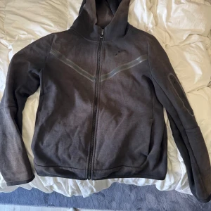 Svart Nike hoodiejacka med dragkedja - Säljer min svarta Nike tech storlek 160-170 säljes då jag har 2 st såklart är den äkta bilder kan fixas privat på den inga hål ett fel finns på fickan på armen men annars funkar den helt perfekt kan byta mot nån annan hoodie kan skickas men då står köpare för frakt.