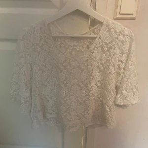 Vit spetsblus med blommönster - Superfin vit blus i spets med blommigt mönster och halvtransparent look. Modellen är croppad med korta ärmar och rund halsringning. Perfekt för dig som vill ha en romantisk och trendig vibe.