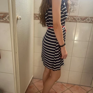 Randig marinblå kortklänning med bälte - Superfin marinblå och vit randig kortklänning från Kids by Kappahl. Klänningen har ett rosa knytbälte i midjan, rund halsringning och korta ärmar med lätt volang. Perfekt för sommarens alla dagar!