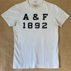 Vit t-shirt Abercrombie & Fitch A&F 1892 - Vit t-shirt från Abercrombie & Fitch med svart tryck 'A&F 1892' på bröstet. Klassisk rund hals och korta ärmar. Modellen är rak och tillverkad i mjuk bomull, perfekt för en clean och sportig look. Använd ett fåtal gånger.