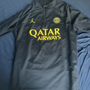 Svart PSG fotbollströja Jordan x Qatar - Svart långärmad PSG fotbollströja med gul logga och detaljer.  Dragkedja är sönder, så den är ihop sydd, så därför kan man inte öpppna den men det funkar ändå 