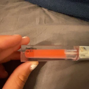 BeautyAct Dew Drop Lip Oil  - Säljer en Dew Drop Lip Oil från BeautyAct. Läppoljan har en snygg röd nyans och kommer i en genomskinlig fyrkantig tub med silvrigt lock. Perfekt för att ge läpparna glans och fukt med en hint av färg.