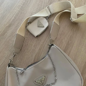 Beige axelväska från Prada med kedja - Säljer en beige axelväska från Prada med bred axelrem och silverfärgad kedjedetalj. Väskan har en liten matchande miniväska fäst på remmen och klassisk Prada-logga framtill. Perfekt för dig som vill ha en stilren och trendig accessoar.