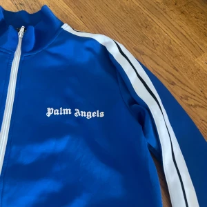 Blå track jacket från Palm Angels - Snygg blå track jacket från Palm Angels med vita ränder längs ärmarna och vit dragkedja. Jackan har hög krage och logga på bröstet. Perfekt för en sportig och trendig look.