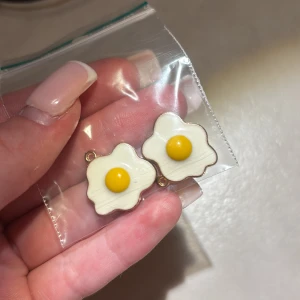 Två berlocker med stekt ägg-design - Superroliga örhänges berlocker formade som stekta ägg med vit och gul färg. De har en vågig kant och en blank gul 'äggula' i mitten. Baksidan är guldfärgad och har en räfflad yta. Perfekta för dig som vill sticka ut med något unikt och lekfullt! 