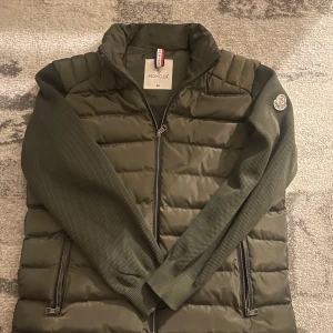 Olivgrön pufferjacka från Moncler - Säljer en olivgrön pufferjacka från Moncler med stickade ärmar och klassisk Moncler-logga på armen. Jackan har dragkedja framtill, två fickor med dragkedja och hög krage. Perfekt för dig som vill ha en snygg och sportig look med exklusiv känsla. Pris kan diskuteras vid snabb affär! Använd 1-2 gånger passade inte mig. (Helt ny)
