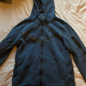 Blå Nike hoodie med dragkedja - Mörkblå hoodie från Nike med hel dragkedja och huva. Jackan har en sportig look med svarta detaljer över bröstet och en ficka med dragkedja på ärmen. Perfekt för chill dagar eller när du vill ha en avslappnad stil. Storlek M använd fåtal gånger 