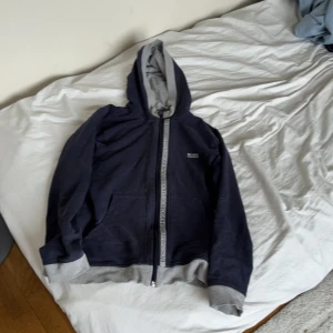 Mörkblå hoodie från Hugo Boss - Snygg mörkblå hoodie från Hugo Boss med gråa muddar och huva. Dragkedja framtill med logga längs kanten och broderad Boss-logga på bröstet. Perfekt för dig som gillar stilrena och bekväma plagg. Materialet är mjukt och känns skönt mot huden.