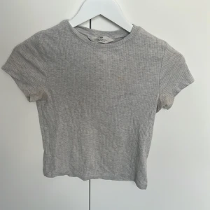 Grå ribbad croppad topp H&M - Grå croppad topp från H&M med ribbad struktur och korta ärmar. Enkel modell med rund halsringning, perfekt till jeans eller kjol. Mjuk och stretchig bomullsblandning som sitter skönt på kroppen. Lite fläckig därför det billiga priset