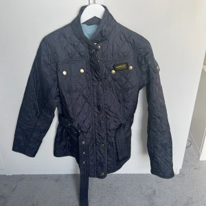 Mörkblå quiltad jacka från Barbour - Quiltad mörkblå jacka från Barbour International med bälte i midjan, guldiga knappar och dragkedja. Jackan har lång ärm, hög krage och flera fickor framtill. Insidan är ljusblå och materialet är lätt och syntetiskt, perfekt för höst och vår. Pris går diskutera!💓