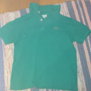 Grön pikétröja från Lacoste - Snygg grön pikétröja från Lacoste med klassisk krage och två knappar framtill. Ikoniska krokodilloggan på bröstet. Kortärmad och tillverkad i bomull, perfekt för en chill och sportig stil. Är barnstorleken 140