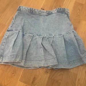 Ljusblå volangkjol i jeans - Supersöt ljusblå jeanskjol med volangkant och hög midja. Kjolen har dragkedja i sidan och knappstängning i midjan. Perfekt för dig som gillar en chill och trendig stil. Passar till sneakers eller boots för en cool look.