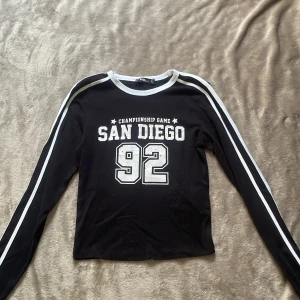 Svart långärmad San Diego-tröja från NEW YORKER  - Svart långärmad tröja från FB Siser med vit text 'San Diego 92' och sportig collegekänsla. Vita ränder längs ärmarna och vit ribbad kant vid halsen. Perfekt för dig som gillar en avslappnad och trendig stil plus helt oanvänd (bara provad).