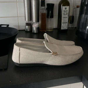 Beige loafers med gulddetalj - Stilrena beige loafers med diskret gulddetalj vid sidan. Skorna har en platt sula och är tillverkade i ett tygmaterial med lätt struktur. Perfekta för en clean och avslappnad look. Knappast använda, nypris: 950 
