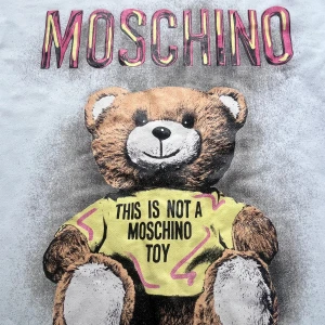 Vit Moschino tröja med björntryck - Säljer en vit långärmad tröja från Moschino med ikoniskt björntryck och texten 'This is not a Moschino toy' på framsidan. Tröjan har rund halsringning och färgglada detaljer i trycket. Perfekt statement piece för dig som gillar streetstyle och coola prints.