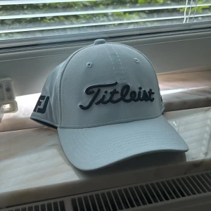 Vit keps från Titleist - Snygg vit keps från Titleist med broderad svart logga framtill och detaljer på sidorna. Klassisk böjd skärm och justerbar passform. Perfekt för golfbanan eller en sportig look.