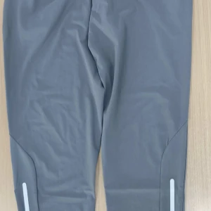 Grå träningsbyxor från Nike - Snygga grå träningsbyxor från Nike med smal passform och diskret logga på höften. Byxorna har reflekterande detaljer vid bensluten och är tillverkade i ett stretchigt syntetmaterial som passar perfekt för träning eller löpning.helt oanvänd möts upp i goteborg!