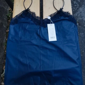 Mörkblå linne med spetsdetaljer - Supersnygg mörkblå linne från Mandy med tunna axelband och spets längs urringningen. Gjord i ett glansigt material som ger en lyxig känsla. Perfekt att styla med jeans eller kjol för en trendig look.