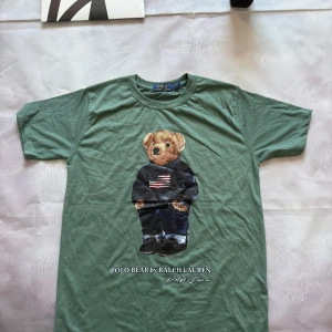 Grön Polo Bear t-shirt från Ralph Lauren - Snygg grön t-shirt från Polo Ralph Lauren med ikoniska Polo Bear-trycket framtill. Klassisk rund hals och korta ärmar. Tillverkad i mjuk bomull som känns skön mot huden. Perfekt för dig som gillar streetstyle och vill sticka ut med en cool printad tee.