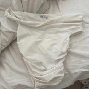Vit offshoulder topp från Zara - Säljer en snygg vit offshoulder topp från Zara i storlek S. Toppen har en draperad design. Perfekt att styla med jeans eller kjol för en trendig look.