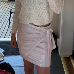 Glittrig omlottkjol i ljusrosa - Säljer en superfin ljusrosa glittrig omlottkjol med knyt i sidan. Kjolen har en kort passform och skimrar snyggt i ljuset – perfekt för dig som vill sticka ut lite extra.