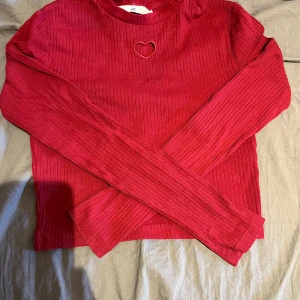 Röd ribbad långärmad tröja med hjärtformad detalj från H&M - Säljer en röd ribbad långärmad tröja från H&M med en söt hjärtformad cut-out vid halsen. Tröjan är croppad och har en tight passform, perfekt för att ge en trendig look.