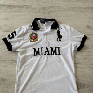 Ralph Lauren piké - En helt ny Vit piké t-shirt från Polo Ralph Lauren med svart krage och detaljer. Stor broderad logga på bröstet, MIAMI-tryck och emblem på ena sidan. Klassisk och sportig stil, perfekt för en avslappnad look. Perfekt inför sommaren! Hör av dig vid minsta fundering!☺️👍🏼