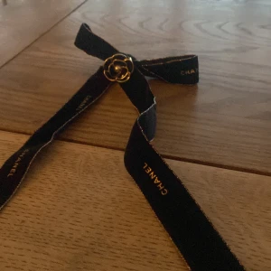 Svart rosett från Chanel  - Säljer en svart rosett från Chanel med en elegant guldfärgad blomdetalj framtill. Bandet har diskret Chanel-logga i guld längs kanten. Perfekt för att lyfta vilken outfit som helst. 
