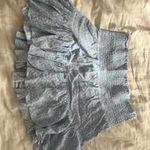 Blå blommig volangkjol - Supersöt blå kortkjol med små vita blommor och volangkant. Kjolen har smock i midjan som gör den bekväm och sitter snyggt. Perfekt för varma dagar och enkel att styla med en crop top eller hoodie.