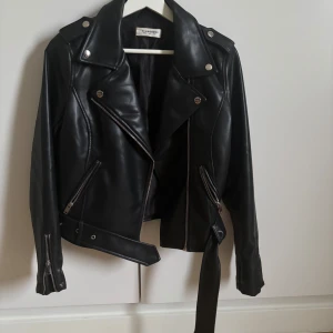 Svart skinnjacka från Glamorous - Säljer en svart skinnjacka från Glamorous med klassisk bikerstil. Jackan har silvriga dragkedjor, knappar och ett bälte i midjan. Snyggt snitt med långa ärmar och bred krage. Skriv för fler bilder🩷