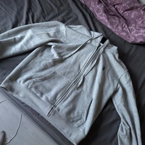 Grå hoodie från Polo Ralph Lauren - Säljer en grå hoodie från Polo Ralph Lauren i storlek M. Tröjan har dragkedja, huva med snören och två fickor framtill. Klassisk broderad logga på bröstet och ribbade muddar vid ärmslut och nederkant.