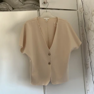 Beige blus med kort ärm och knappar - Säljer en stilren beige blus med v-ringning och två dekorativa knappar framtill. Blusen har korta ärmar och en lös passform, perfekt för en enkel och snygg look. Produkten är i nyskick och har aldrig använts